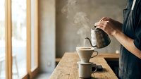 Man pouring hot coffee from metal pot into white cup on wooden table with steam rising in cozy kitchen morning light atmosphere for cafe lifestyle concepts.