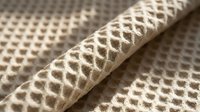 Closeup of woven beige and cream textured fabric with diamond lattice pattern, showing rolled edge detail for interior design and textile projects.