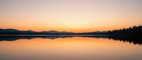 Serene lake at sunrise with mirror-like water reflecting golden sky, distant mountains and evergreen forest silhouettes creating tranquil nature landscape for backgrounds and meditation content.