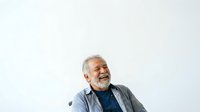 Senior man with gray hair and beard, wearing casual blue shirt and gray cardigan, smiling while looking upward against white background. Isolated portrait for design use.