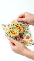 Hands holding healthy vegetable wrap with colorful peppers and greens in floral eco-friendly paper on white background for lunch meal preparation.