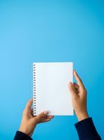 Hands holding blank spiral notebook against bright blue background, ready for writing or note-taking projects.