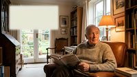Senior Caucasian man reading newspaper in cozy home library with warm lighting, bookshelves, and comfortable armchair creating peaceful retirement lifestyle.