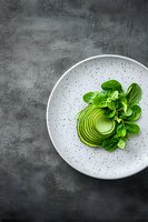 Sliced avocado arranged in spiral pattern on speckled white plate with fresh corn salad greens, shot from above on dark concrete background.