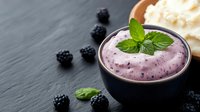 Creamy blackberry yogurt mousse garnished with fresh mint leaves in dark ceramic bowl on slate surface, scattered berries and whipped cream for dessert serving.