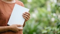 Person in terracotta shirt holding blank white card outdoors with green blurred background, suitable for mockups, advertising, and promotional campaigns.