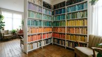 Cozy home library corner with color-coordinated books arranged on floor-to-ceiling shelves, creating rainbow gradient effect in modern reading room.