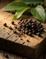 Black peppercorns scattered on rustic wooden cutting board with fresh green leaves for culinary seasoning and cooking applications.