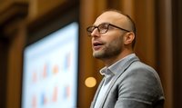 Confident bald Caucasian businessman with glasses and grey jacket speaking at conference presentation, side view profile against blurred projection screen background.
