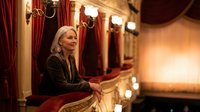 Elegant senior woman with gray hair in black attire sitting in ornate theater box with red velvet curtains and golden details during evening performance.
