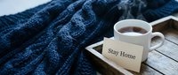 Steaming cup of hot tea on a wooden tray with a Stay Home note card and cozy blue knitted blanket in the background.