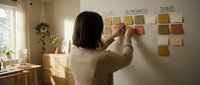 Woman with dark hair wearing a beige sweater organizing sticky notes on a wall with task management labels. Bright natural light from window illuminates workspace.