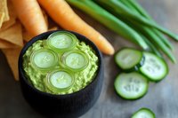 Fresh avocado dip garnished with cucumber slices in black bowl, served with raw carrots and green onions on concrete background, healthy appetizer.