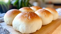 Fresh homemade dinner rolls with golden brown tops sprinkled with salt, arranged on wooden cutting board. Soft, fluffy texture shows through light surface.