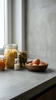 Minimalist kitchen counter with glass jars containing pasta, pickles, beans and wooden bowl of fresh eggs near window for healthy cooking and food storage.
