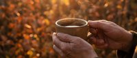Hands holding steaming hot cup of coffee or tea outdoors during golden autumn sunset with warm bokeh lights creating cozy atmospheric moment for relaxation.