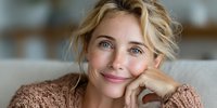 Young blonde Caucasian woman with curly hair smiling warmly while resting chin on hand in cozy knit sweater at home. Natural lifestyle portrait for wellness content.