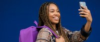 Young African American woman with braids taking selfie with smartphone while carrying purple backpack against blue background for social media content.