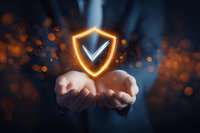 Glowing shield with checkmark hovering over open hands against dark background with orange light particles, symbolizing digital protection and security.