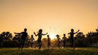 Group of children playing together outdoors in a grassy field during golden hour sunset, running and jumping with arms raised in joy and celebration.