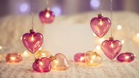 Pink heart-shaped glass ornaments hanging with warm fairy lights creating romantic Valentine's Day decoration on soft textured surface.