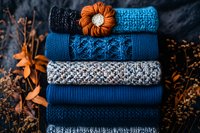 Stack of folded blue knitted sweaters with orange flower brooch, surrounded by autumn dry branches on dark background.