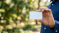 Male professional with medium skin tone holding blank white business card outdoors with blurred green foliage background, suitable for corporate branding and networking materials.