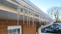 Glistening icicles hanging from white house gutter against winter sky, showing seasonal weather conditions and frozen water formations on residential building.