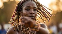 Young African woman with dreadlocks pointing directly at camera with confident expression during golden hour sunset light creating warm atmospheric portrait.