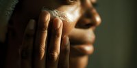 Close-up of person with dark skin touching their face, illuminated by soft, dramatic side lighting that highlights facial contours and texture of skin.