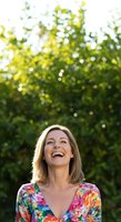 Caucasian woman with shoulder-length blonde hair, smiling upward outdoors with vibrant green foliage and bokeh sunlight in background. Wearing colorful floral print top.