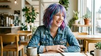 Young Caucasian woman with colorful purple hair laughing joyfully in a cozy cafe, sitting at a table with a notebook and coffee mug.