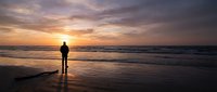 Silhouette of man standing on wet beach at sunset, watching ocean waves and dramatic sky with golden and purple clouds reflecting on sand.