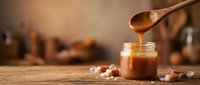 Golden caramel sauce dripping from wooden spoon into glass jar on rustic kitchen counter with warm lighting for culinary and dessert recipes.