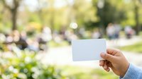 Hand holding blank white card in sunny park with blurred green trees and people in background, suitable for business messaging, promotional campaigns, and outdoor advertising concepts.