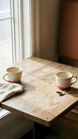 Two ceramic coffee cups on rustic wooden table by window with warm natural light creating cozy morning atmosphere for cafe or home interior design.