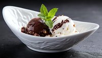 Chocolate and vanilla ice cream scoops garnished with fresh mint leaves in elegant white ceramic bowl against dark background, drizzled with chocolate sauce.