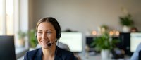 Professional female customer service representative with headset smiling in modern office environment with computers and plants in background.
