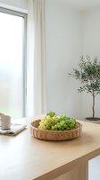 Fresh green and yellow grapes in woven basket on wooden table by bright window with white curtains, potted olive plant, and coffee cup for home decor styling.
