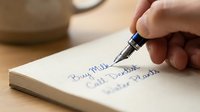 Hand writing daily to-do list in notebook with fountain pen on wooden desk for productivity and organization planning.
