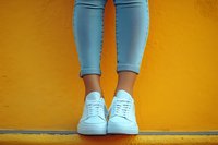 Female legs in light blue skinny jeans and white sneakers standing against vibrant yellow wall, modern casual urban fashion style for youth culture and lifestyle.
