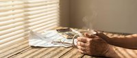 Elderly person's hands resting on wooden table with cigarette and ashtray, sunlight streaming through blinds, depicting smoking habit and health concerns.