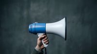 Hand holding blue and white megaphone against dark textured wall background for communication, announcement, marketing and promotional campaigns.