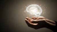 Glowing brain hologram floating above open hands with sparkling light effects on dark background representing artificial intelligence and mental power.