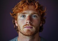 Close up portrait of young redhead man with freckles and blue eyes looking at camera against dark background. Natural facial expression and curly ginger hair.