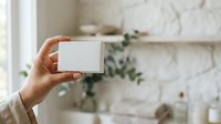 Cropped hand holding blank white card against blurred interior background with marble wall and potted plant, suitable for branding, business card mockups, and promotional designs.