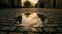 Old stone street with puddle reflecting golden sunset sky and historic architecture, suitable for travel, nostalgia, and urban design projects.