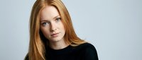 Young redhead woman with long straight hair wearing black sweater looking at camera with serious expression against neutral background for portrait photography.