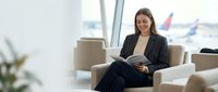 Young professional businesswoman in dark suit reading documents while sitting in modern airport lounge with planes visible through windows.