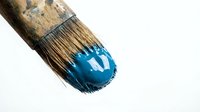 Paintbrush with worn wooden handle applying bright blue paint stroke on white surface for art and design projects.